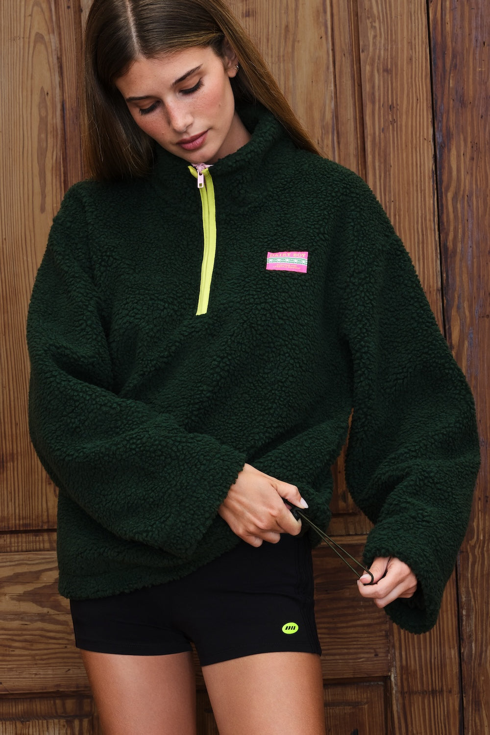A woman wearing a dark forest green sherpa fleece pullover with a bright neon yellow zipper and a small pink logo patch on the chest, paired with black athletic shorts, standing against a rustic wooden plank wall.
|M1