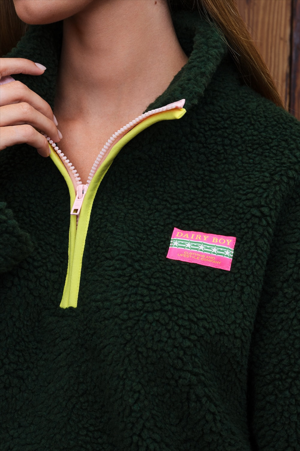 Close-up of a person wearing a dark green sherpa fleece quarter-zip pullover with a bright yellow zipper trim and a pink patch label on the chest.
|M1