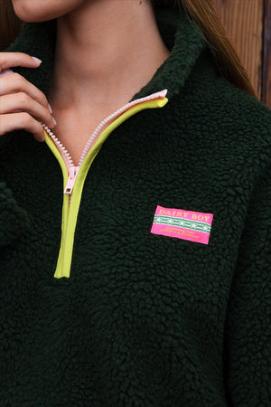 Close-up of a person wearing a dark green sherpa fleece quarter-zip pullover with a bright yellow zipper trim and a pink patch label on the chest.
|M1