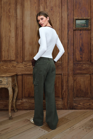 A woman stands in a rustic room with wooden paneling, wearing a fitted white long-sleeve top and dark olive green wide-leg cargo pants, shown from the back.
|M1