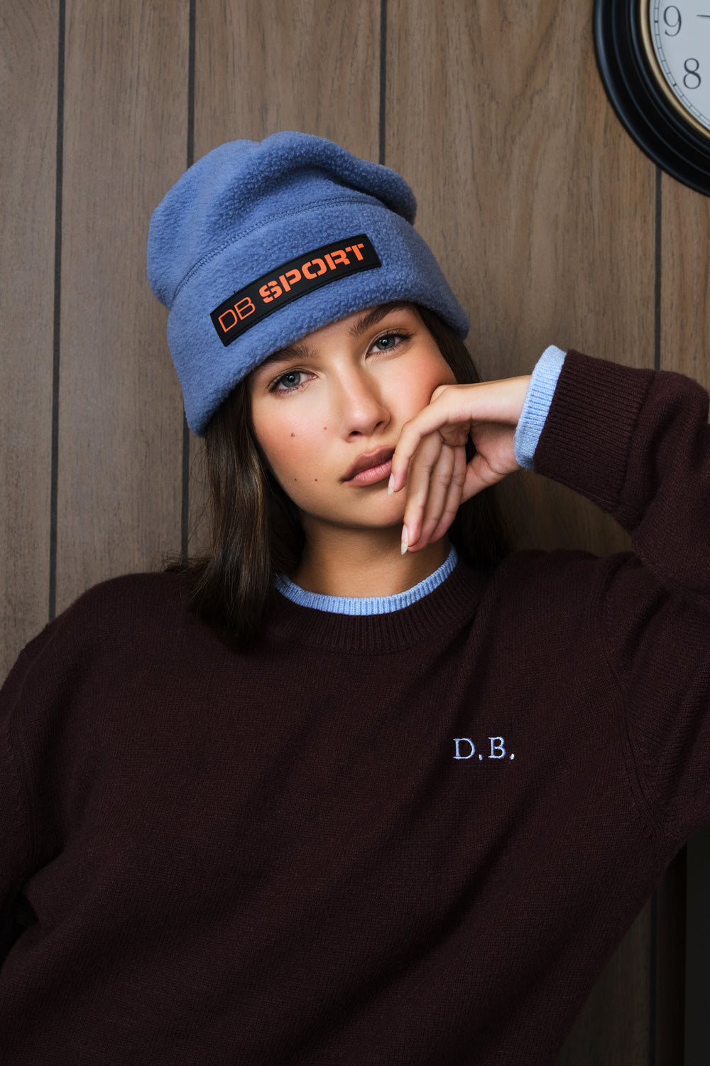A woman wearing a dusty blue knit beanie with an orange and black 'DB SPORT' patch and a dark brown sweater with a light blue collar, posed against a wooden wall.