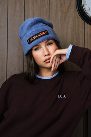 A woman wearing a dusty blue knit beanie with an orange and black 'DB SPORT' patch and a dark brown sweater with a light blue collar, posed against a wooden wall.