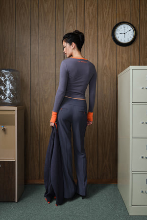A woman stands with her back to the camera in an office-like room with wood-paneled walls, holding a dark grey outfit. She is wearing a fitted long-sleeve top and matching pants with bright orange cuffs and waist detail.
|M1