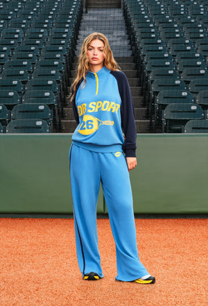 A woman stands on a baseball field wearing a light blue and navy athletic tracksuit with yellow accents. The background shows rows of empty stadium seats.
|M1
