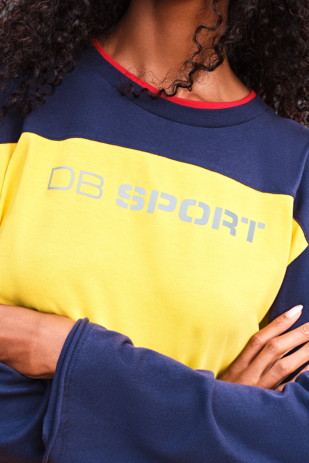 A close-up of a person wearing a colorblock shirt with navy blue sleeves and shoulders, a bright yellow chest panel, and a red inner collar trim. The yellow panel features the text 'DB SPORT' in gray.
|M1