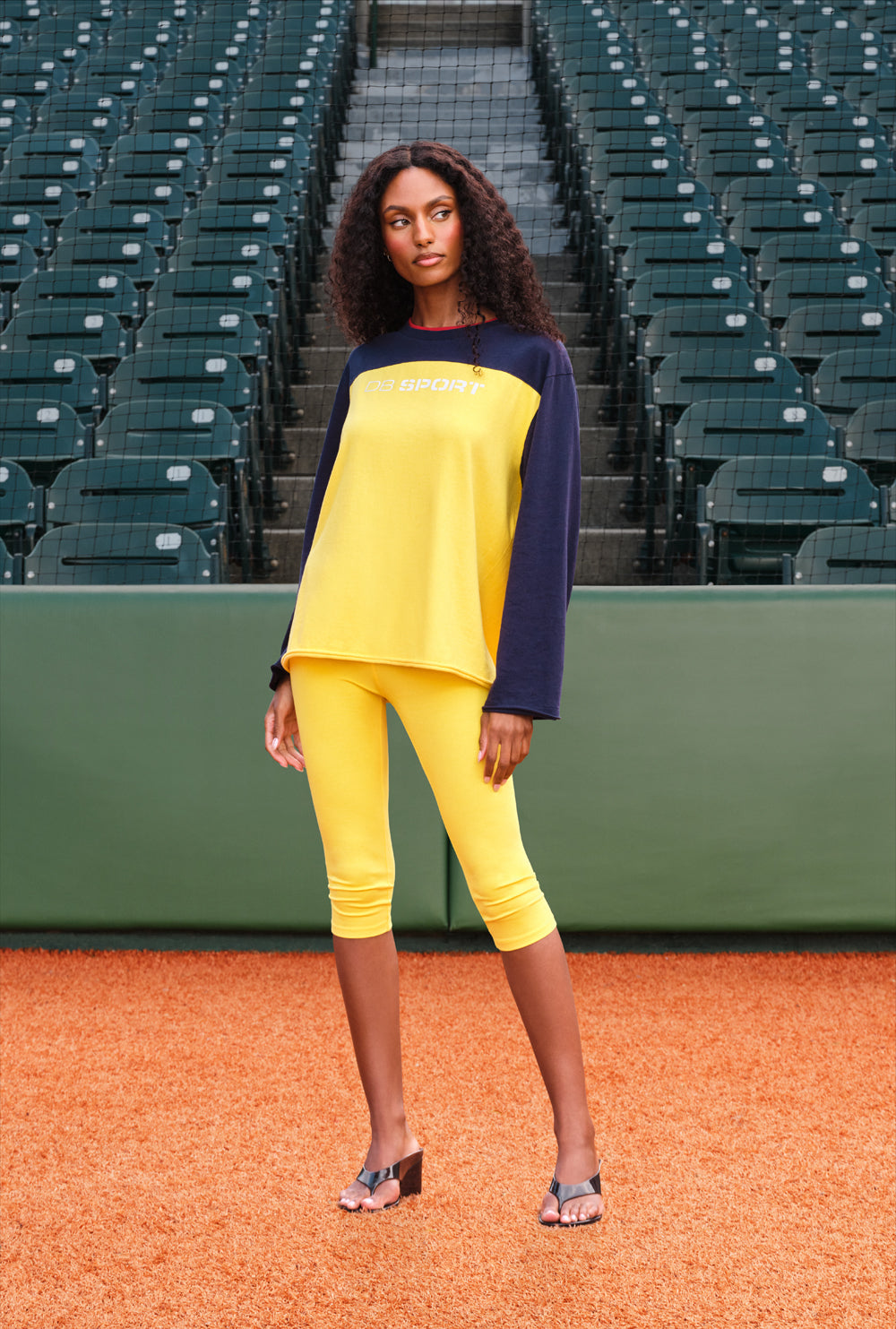 A woman stands on a baseball field wearing a bright yellow and navy long-sleeve top with matching yellow capri leggings. The background shows rows of green stadium seats.
|M1