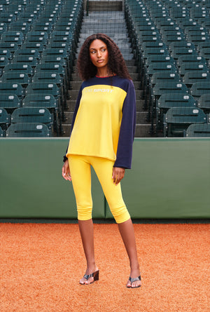 A woman stands on a baseball field wearing a bright yellow and navy long-sleeve top with matching yellow capri leggings. The background shows rows of green stadium seats.
|M1