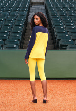 A woman stands on a baseball diamond wearing a colorblock athletic set consisting of a long-sleeve top and matching capri leggings. The top features navy blue sleeves and shoulders, with a bright yellow body.
|M1