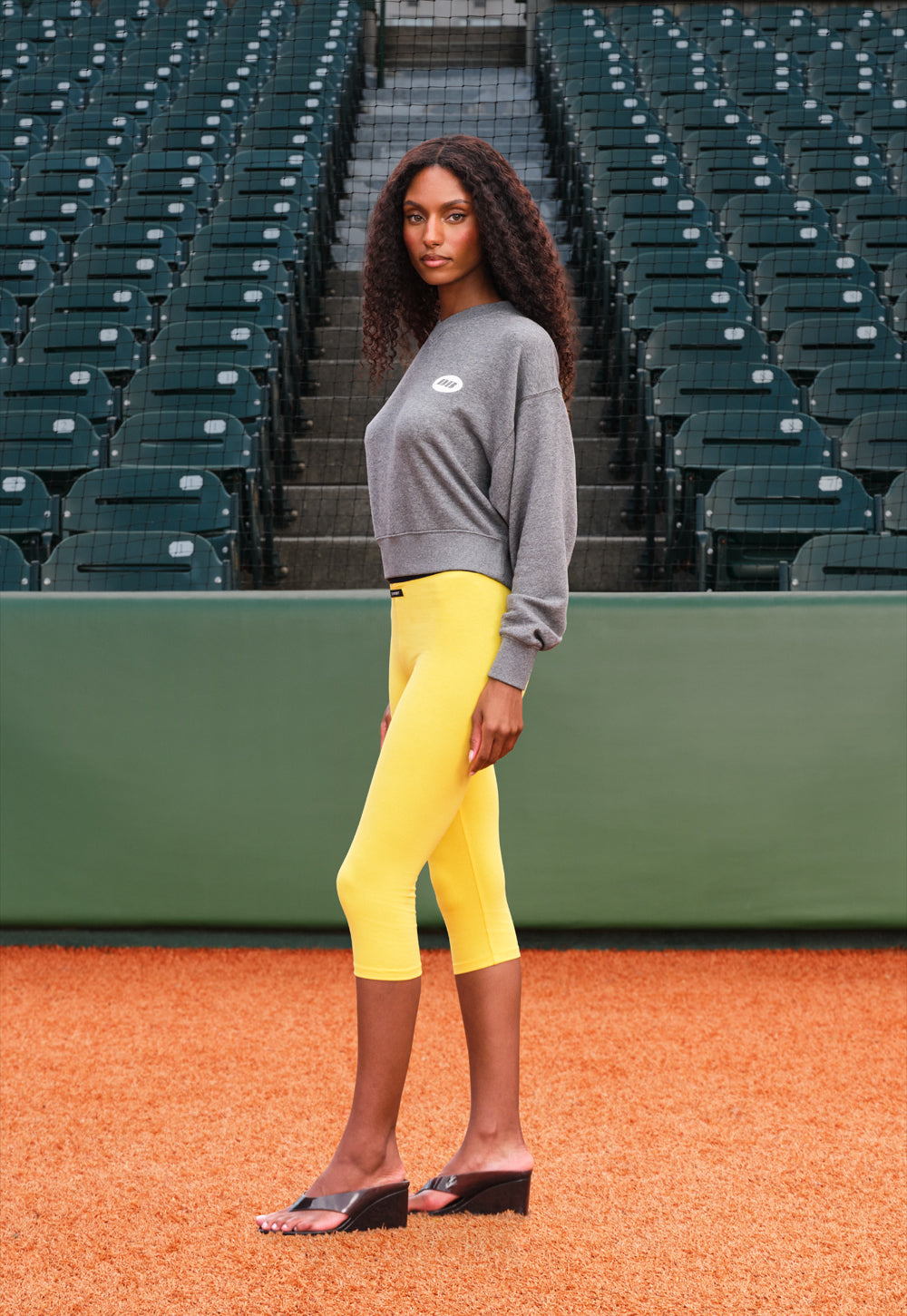 A woman stands on a baseball diamond in front of green stadium seating, wearing a gray sweatshirt, bright yellow cropped leggings, and black wedge sandals.
|M1
