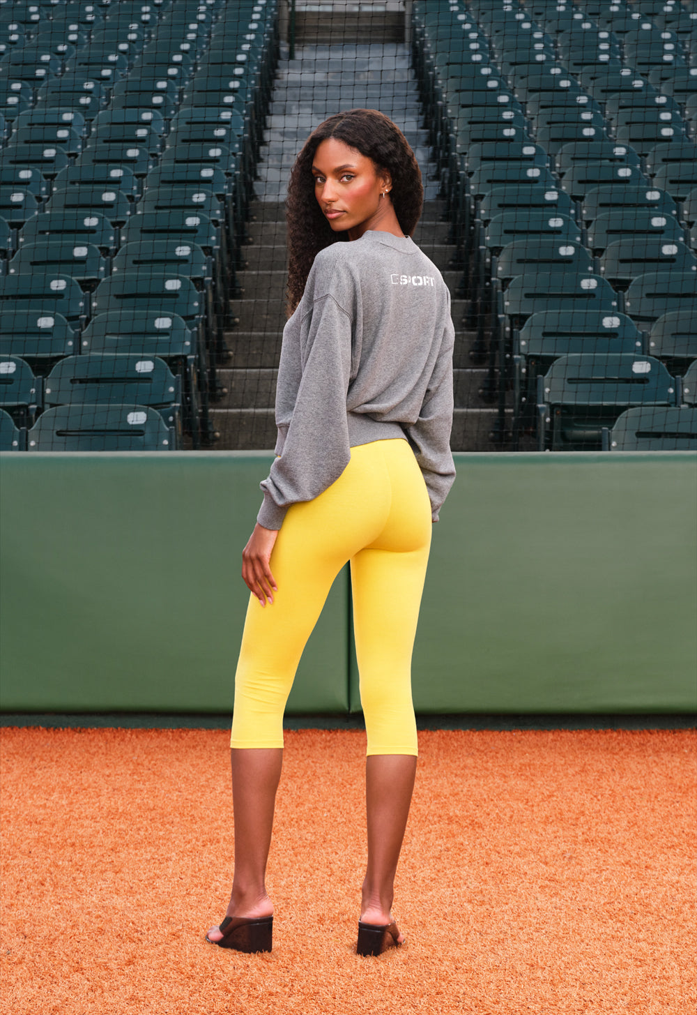 A woman stands on a baseball diamond wearing a gray long-sleeve athletic top and bright yellow fitted capri leggings, photographed from the back. Empty stadium seating and a green wall are in the background.
|M1