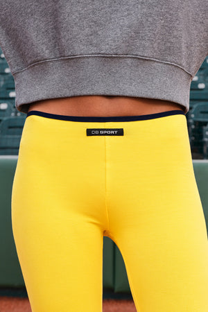 Close-up of a person wearing bright yellow athletic leggings with a black waistband and a small black brand label reading 'DB SPORT' at the center front. The person is also wearing a gray cropped sweatshirt, and the background shows blurred stadium seating.
|M1