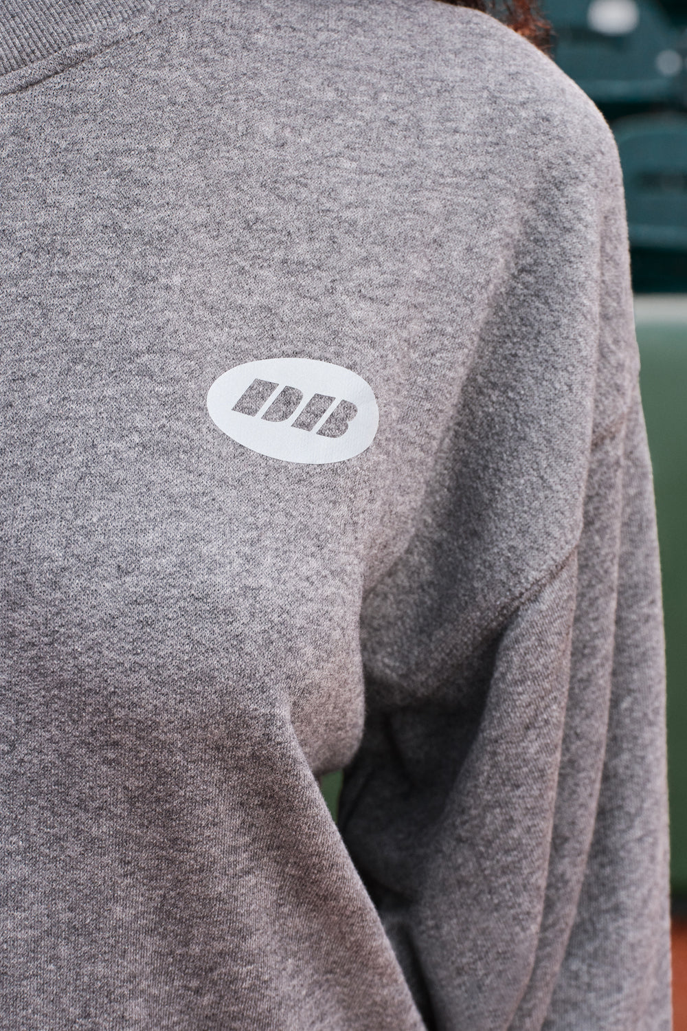 Close-up of a heather gray crewneck sweatshirt featuring a white oval chest patch with the letters 'DB' in white.
|M1