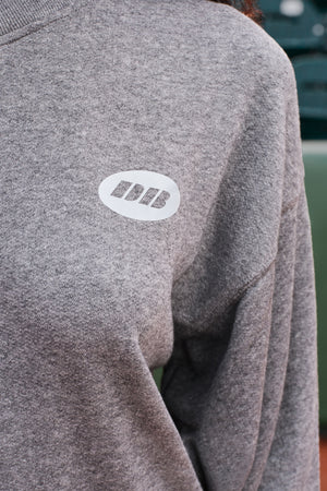 Close-up of a heather gray crewneck sweatshirt featuring a white oval chest patch with the letters 'DB' in white.
|M1
