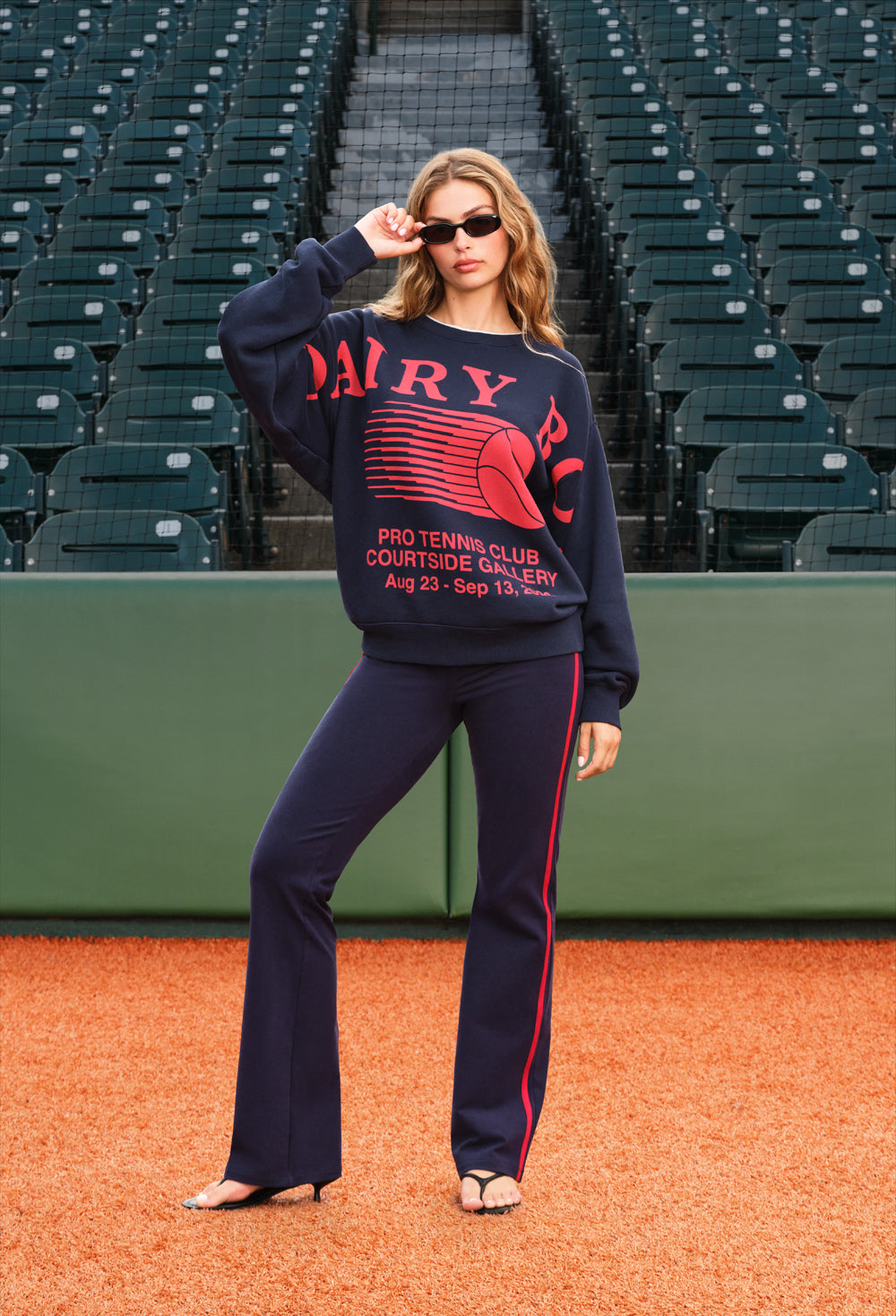 A woman posing on a baseball diamond wearing a navy blue sweatshirt and matching flared pants with red and white graphics. The sweatshirt features 'DAIRY BOY' in red with a tennis ball graphic.
|M1 