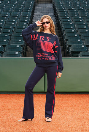 A woman posing on a baseball diamond wearing a navy blue sweatshirt and matching flared pants with red and white graphics. The sweatshirt features 'DAIRY BOY' in red with a tennis ball graphic.
|M1 