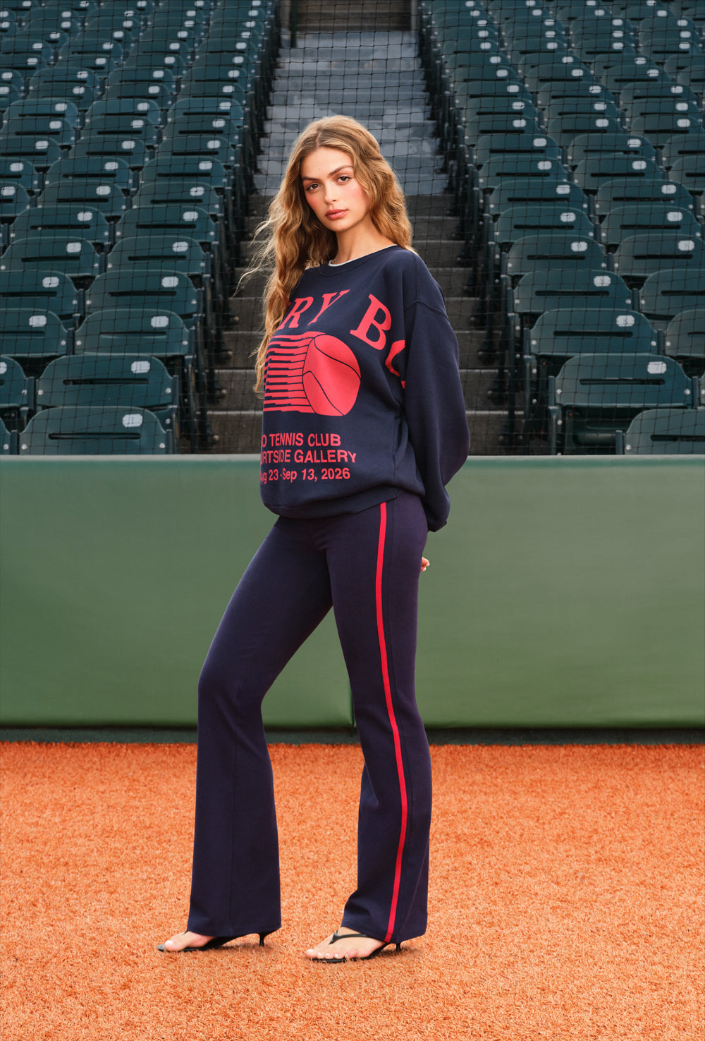 A woman stands on an orange baseball field wearing a navy blue sweatshirt and matching flared pants with a red side stripe and red graphic text. Behind her are rows of dark stadium seats.
|M1