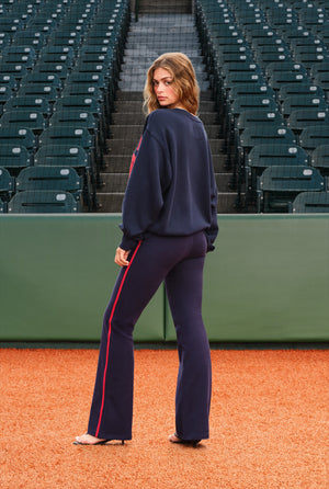 A woman stands on an orange baseball field wearing a navy blue sweatshirt and matching flared pants with a red side stripe, photographed in front of stadium seating.
|M1
