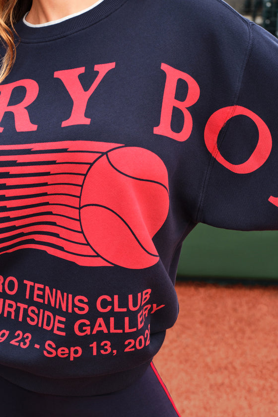 Close-up of a navy blue sweatshirt with a large red graphic of a tennis ball and partial red text, worn by a person on an outdoor court with orange ground visible in the background.
|M1