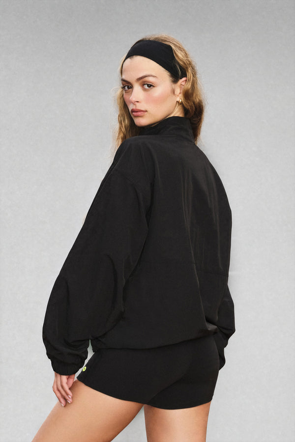A woman wearing a black long-sleeve mock-neck athletic jacket and matching black shorts, photographed from the back against a neutral gray studio background.
|M1