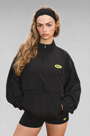 A woman modeling a black half-zip pullover jacket with a small yellow logo on the left chest, paired with black shorts, against a plain gray studio background.
|M1
