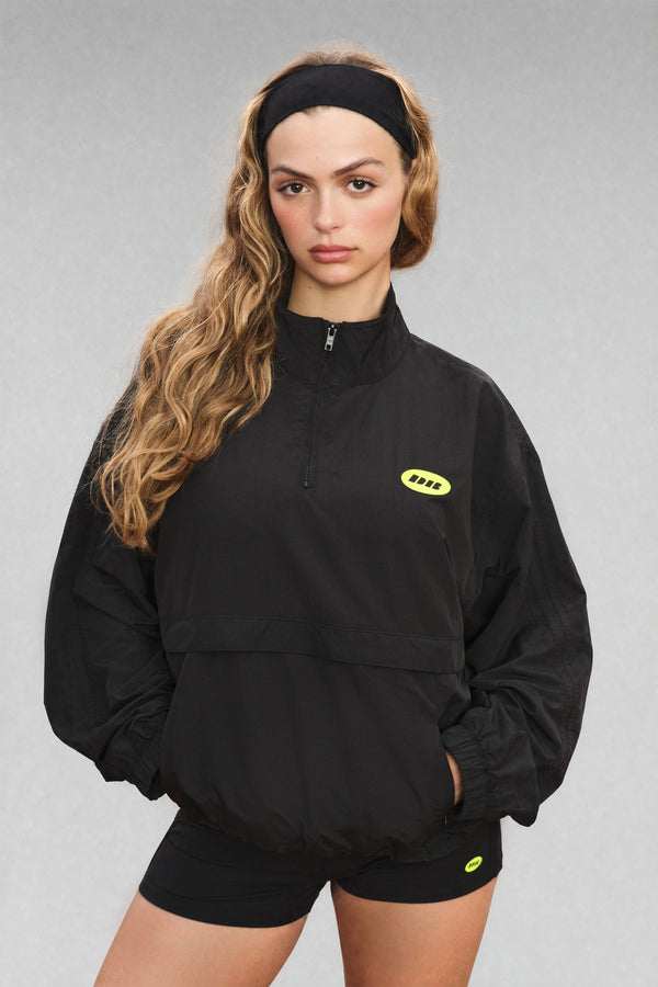 A woman modeling a black half-zip pullover jacket with a small yellow logo on the left chest, paired with black shorts, against a plain gray studio background.
|M1
