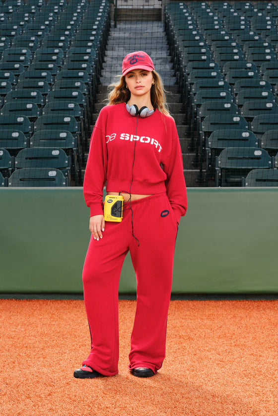 A woman stands on a baseball field wearing a matching red tracksuit with a red cap and headphones around her neck, with stadium seats in the background.
|M1