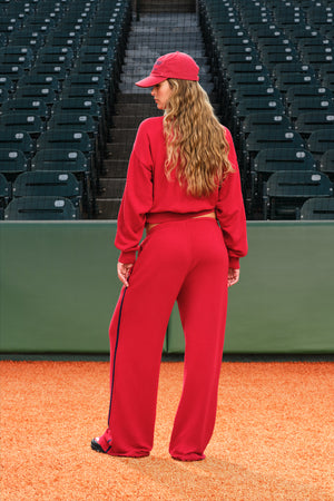 A woman stands on an orange baseball field wearing a matching red tracksuit with a red cap, viewed from behind. The background shows rows of empty dark stadium seats.
|M1