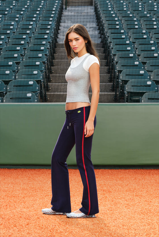 A woman stands on a baseball diamond wearing navy flared athletic pants with a red side stripe, a light gray short-sleeve crop top, and white sneakers. Empty green stadium seats rise in the background.
|M2