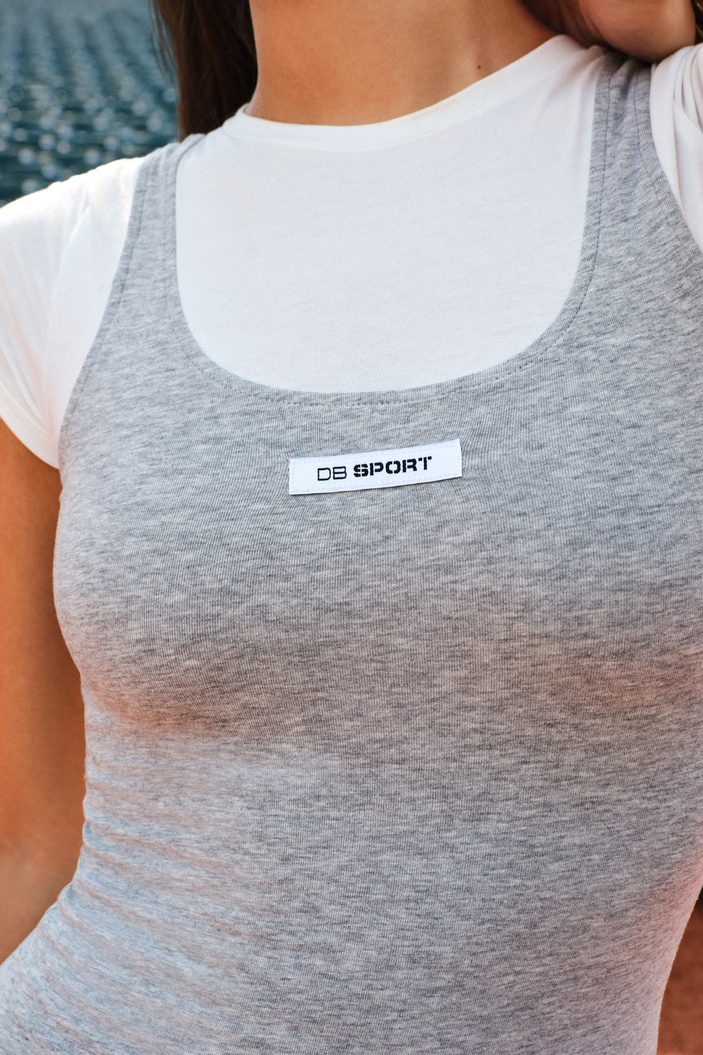 A close-up of a woman wearing a heather gray athletic tank top layered over a white short-sleeve shirt. The gray tank top features a scoop neckline and a white rectangular chest patch with the text 'DB SPORT'.
|M2