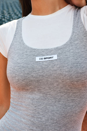 A close-up of a woman wearing a heather gray athletic tank top layered over a white short-sleeve shirt. The gray tank top features a scoop neckline and a white rectangular chest patch with the text 'DB SPORT'.
|M2