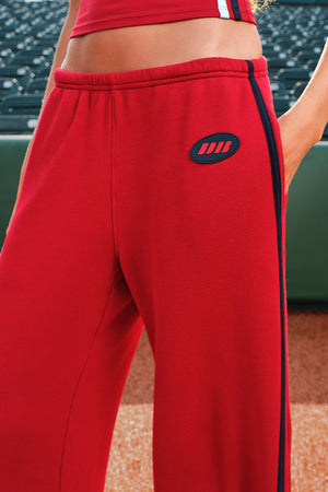Close-up of a person wearing bright red athletic sweatpants with a dark side stripe and a small oval logo patch on the upper thigh. The background shows stadium seating.
|M1