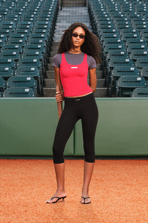 A woman stands on a baseball diamond in front of stadium seating, wearing a bright pink sleeveless tank top over a gray short-sleeve shirt, black capri leggings, and black sandals.
|M1