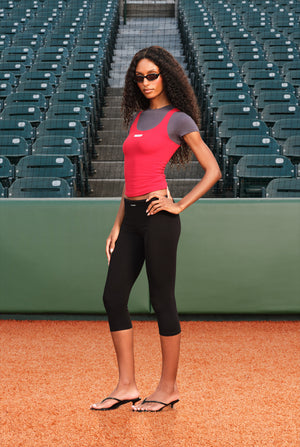 A woman stands on a baseball diamond wearing a pink and gray athletic tank top, black capri leggings, and black flip-flop sandals, with rows of green stadium seats in the background.
|M1