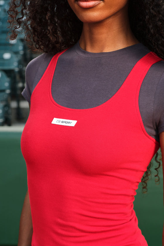 A close-up of a woman wearing a bright red athletic tank top over a dark gray short-sleeve shirt. The red tank top has a scoop neckline and a small white rectangular chest label that reads 'DB SPORT'.
|M1