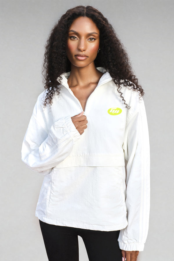 A woman with long, curly dark hair wearing a white zip-up windbreaker jacket with a small lime green logo on the left chest, paired with black pants, against a solid gray studio background.
|M1