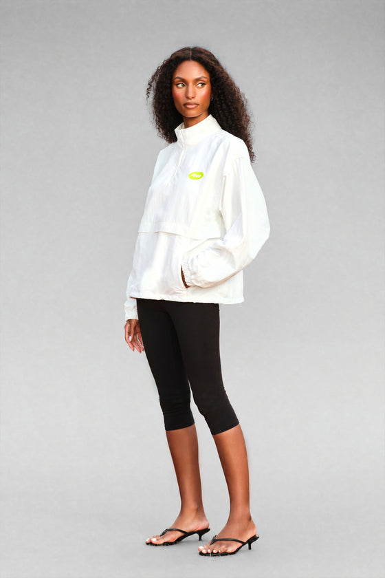 A model wearing a white high-neck pullover jacket with a small lime green chest logo, paired with black capri leggings and black heeled sandals, against a solid gray studio background.
|M1
