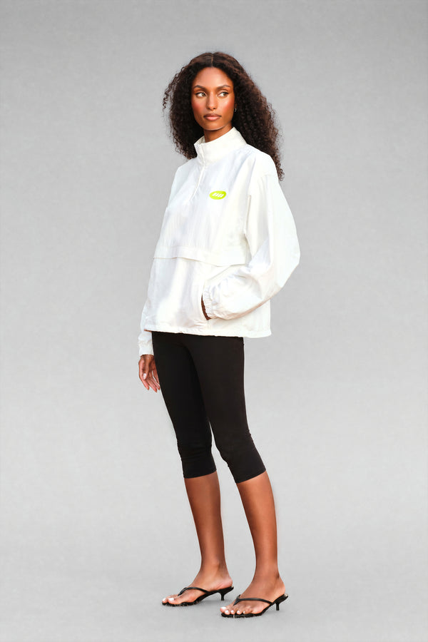 A model wearing a white high-neck pullover jacket with a small lime green chest logo, paired with black capri leggings and black heeled sandals, against a solid gray studio background.
|M1