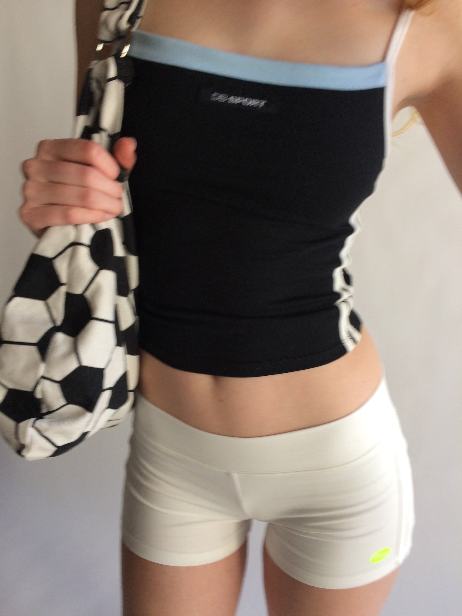 Person wearing a black sports tank and white shorts holding a soccer bag.  |M1