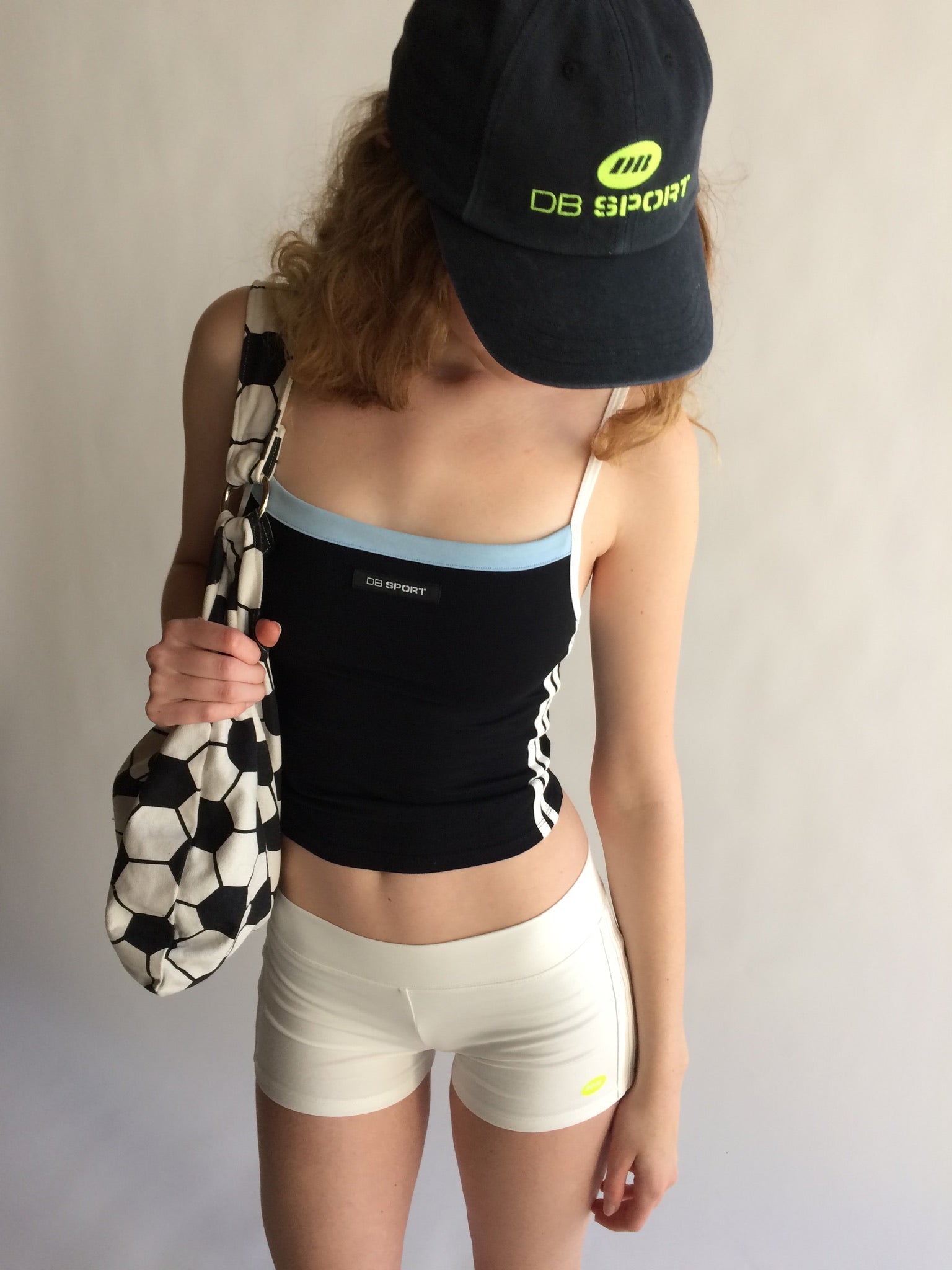 Person wearing a black sports bra and white shorts, holding a soccer ball bag, with a DB Sport cap on a plain background.