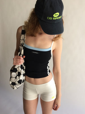 Person wearing a black sports bra and white shorts, holding a soccer ball bag, with a DB Sport cap on a plain background.