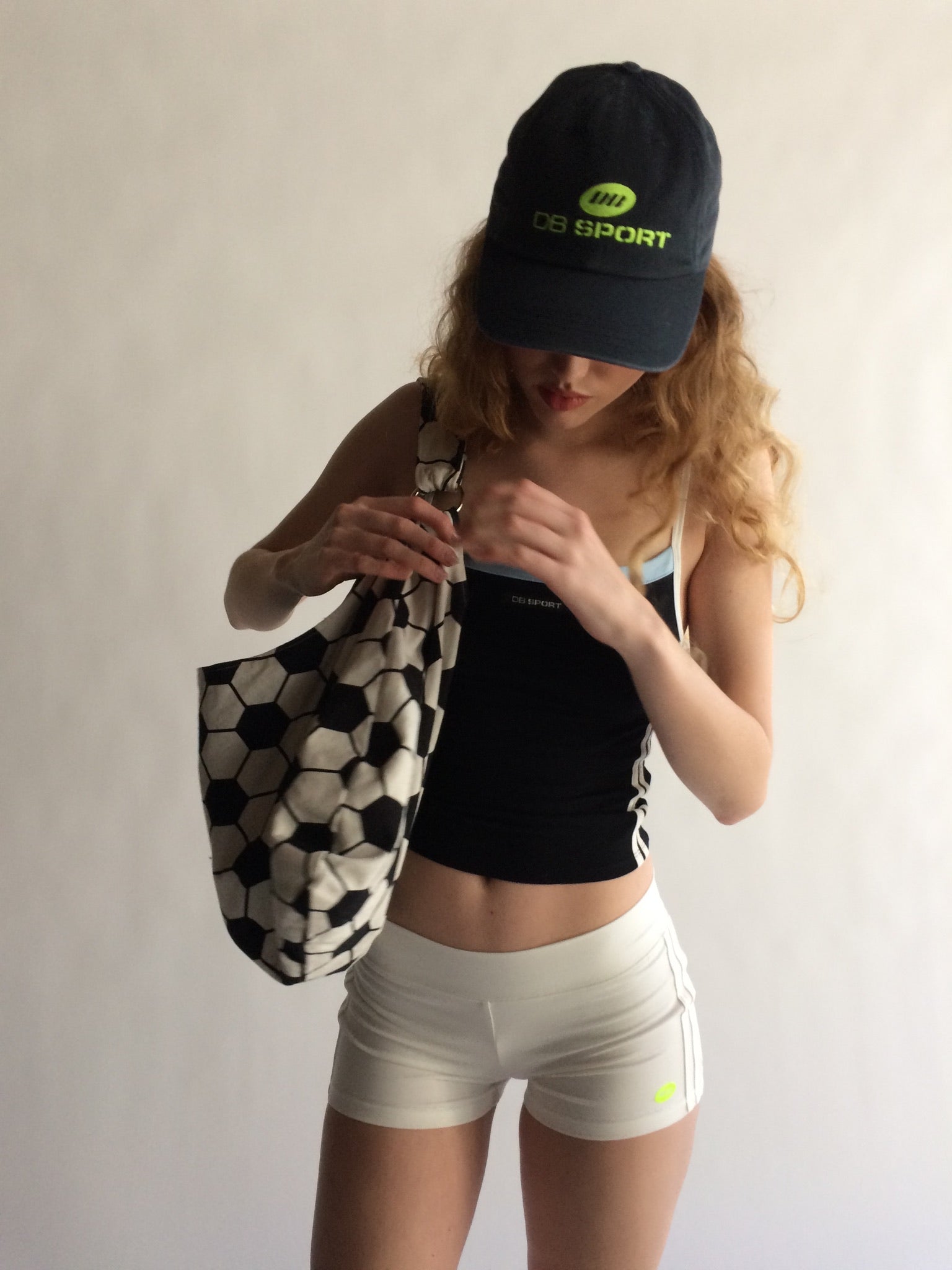 Woman wearing a black cap and sports outfit holding a patterned bag against a plain background