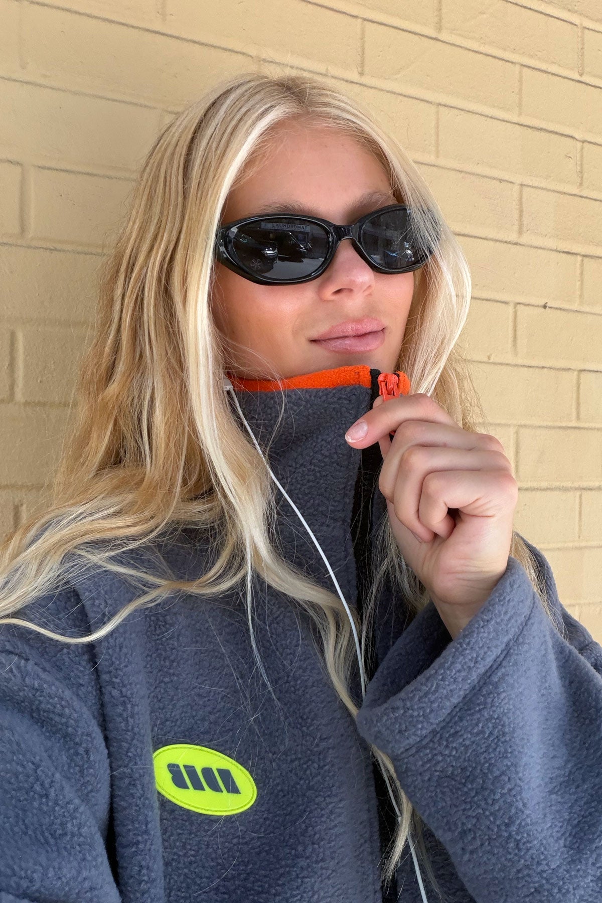 A blonde woman wearing black sunglasses and a dark gray fleece pullover with a bright green logo and orange zipper trim, standing against a beige brick wall.