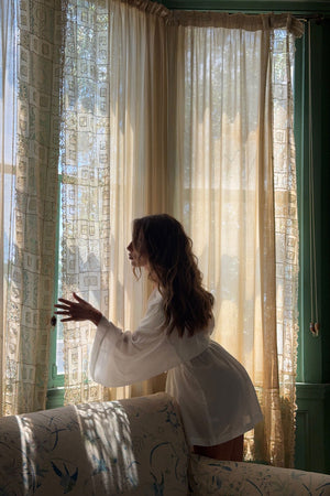 A woman in a white long-sleeve robe stands by a window with sheer cream curtains, looking out while wearing a white robe in a sunlit room.
|M1