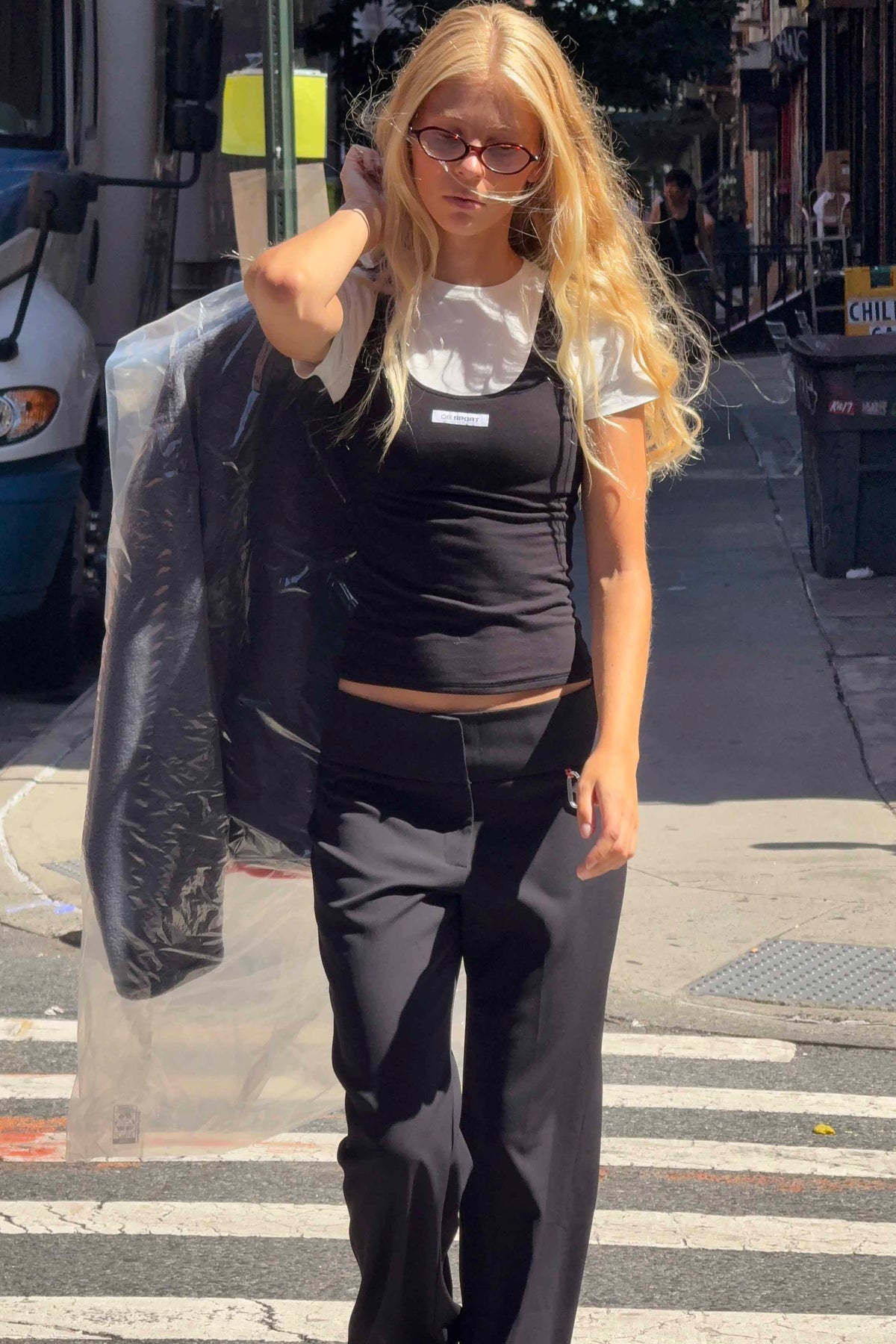 A woman walking on a city street wearing a black sleeveless top, black pants, and a white T-shirt, with a clear plastic garment bag over her shoulder.