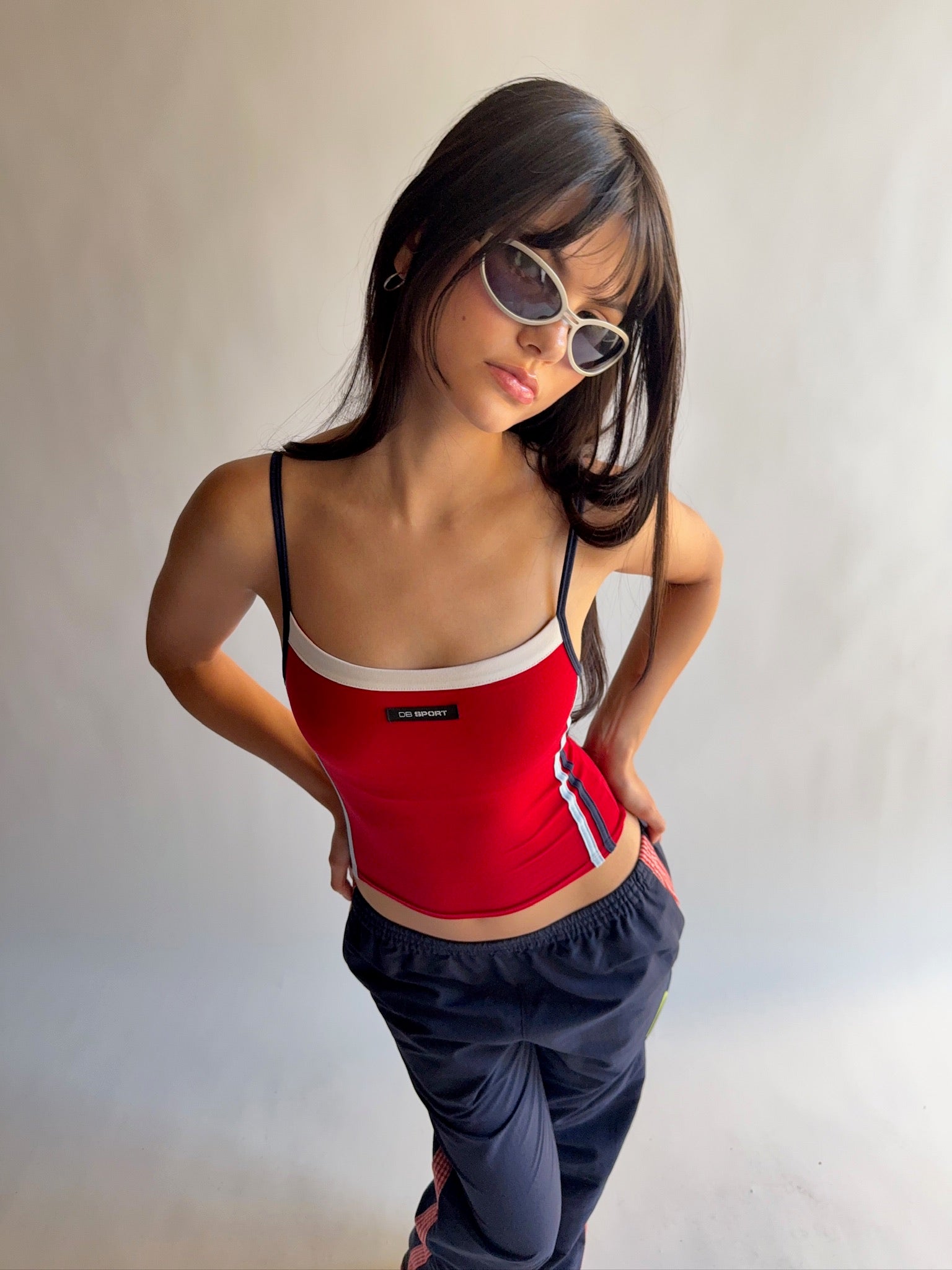 Woman wearing a red tank top and navy pants with sunglasses on a plain background  |M1
