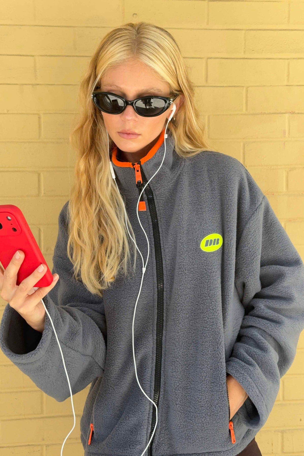 A woman with blonde hair wearing black sunglasses, white earbuds, and a gray fleece jacket with orange accents and a yellow-green circular logo, holding a red smartphone against a yellow brick wall.