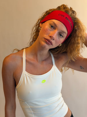Woman wearing a white tank top with a visible brand logo, red headband, and earrings against a plain background. |M1