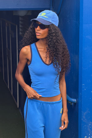 A woman wearing a blue athletic tank top and matching sweatpants with a blue cap and sunglasses, standing in front of a blue wall.
|M1