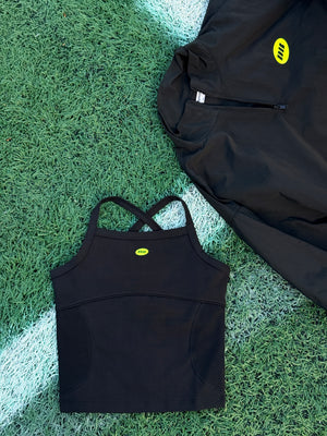 A black athletic zip-up jacket and a matching black sports bra laid flat on green turf with a white line, both featuring a small yellow logo.