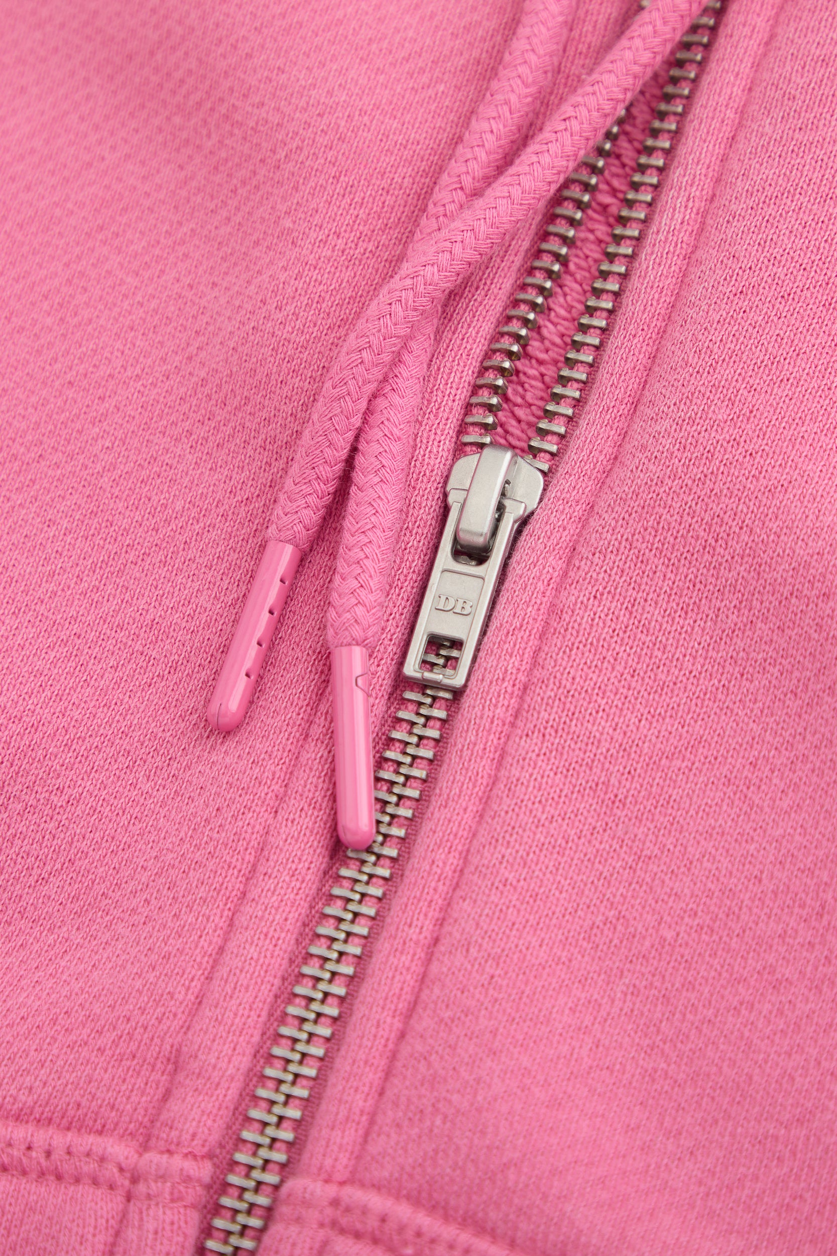 Close-up of the Dairy Boy Strawberry Milk Bottle Zip Up in strawberry pink, highlighting its metal zipper with DB engraved and two matching drawstrings with plastic tips, resting on the soft fabric.